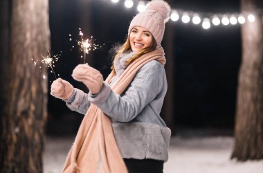 Elinde maytap olan güzel bir kız. Ormanda mutlu kış zamanları. Kız mavi bir ceket giymiş, açık renk bir şapka, atkı ve eldiven takmış. Şenlikli çelenk ışıkları. Noel, yeni yıl.
