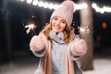 Elinde maytap olan güzel bir kız. Ormanda mutlu kış zamanları. Kız mavi bir ceket giymiş, açık renk bir şapka, atkı ve eldiven takmış. Şenlikli çelenk ışıkları. Noel, yeni yıl.