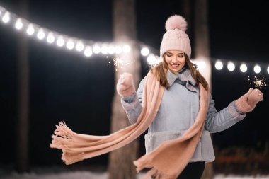 Elinde maytap olan güzel bir kız. Ormanda mutlu kış zamanları. Kız mavi bir ceket giymiş, açık renk bir şapka, atkı ve eldiven takmış. Şenlikli çelenk ışıkları. Noel, yeni yıl.