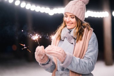 Elinde maytap olan güzel bir kız. Ormanda mutlu kış zamanları. Kız mavi bir ceket giymiş, açık renk bir şapka, atkı ve eldiven takmış. Şenlikli çelenk ışıkları. Noel, yeni yıl.