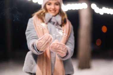 Maytap ellerinde. Ormanda mutlu kış zamanları. Kız mavi bir ceket giymiş, açık renk bir şapka, atkı ve eldiven takmış. Şenlikli çelenk ışıkları. Noel, yeni yıl.