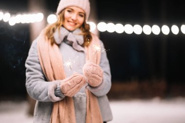 Maytap ellerinde. Ormanda mutlu kış zamanları. Kız mavi bir ceket giymiş, açık renk bir şapka, atkı ve eldiven takmış. Şenlikli çelenk ışıkları. Noel, yeni yıl.