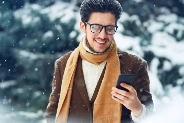 Young man in a coat on the street with a phone. Smiling young man having fun outdoors. Businessman uses a phone.  Man enjoy a winter. Dressed in a coat, sweater and scarf. Winter concept. Snowfall.