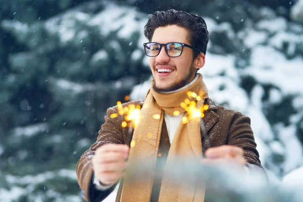 Young man in a coat on the street with sparklers. Smiling young man having fun outdoors. Man enjoy a winter. Dressed in a coat, sweater and scarf. Winter concept. Snowfall.