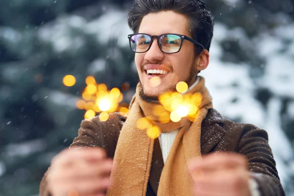 Young man in a coat on the street with sparklers. Smiling young man having fun outdoors. Man enjoy a winter. Dressed in a coat, sweater and scarf. Winter concept. Snowfall.