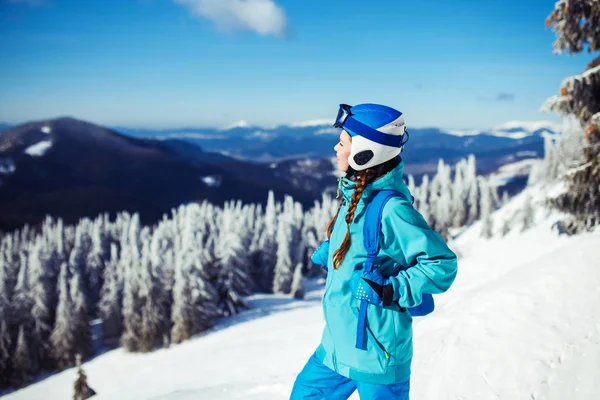A young girl enjoys beauty of winter landscape. A beautiful girl in winter clothes, a blue helmet and jacket is having a great time in the mountains. Concept of travel, leisure, freedom, sport, nature