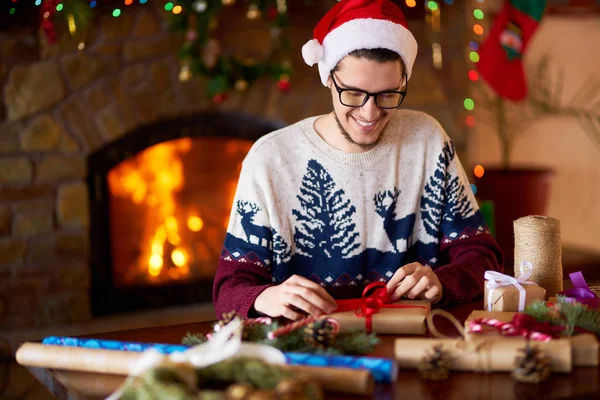 Happy man in a Santa hat creates gifts. The man made them himself and is going to give for the holidays. Concept of gifts and greetings for Christmas and New Year. Beautiful background with fireplace.