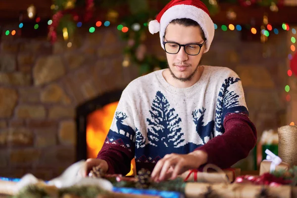 Happy man in a Santa hat creates gifts. The man made them himself and is going to give for the holidays. Concept of gifts and greetings for Christmas and New Year. Beautiful background with fireplace.