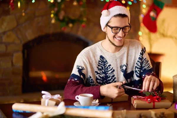 Happy man in a Santa hat creates gifts. The man made them himself and is going to give for the holidays. Concept of gifts and greetings for Christmas and New Year. Beautiful background with fireplace.