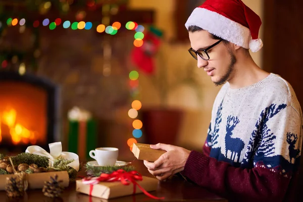 Happy man in a Santa hat creates gifts. The man made them himself and is going to give for the holidays. Concept of gifts and greetings for Christmas and New Year. Beautiful background with fireplace.