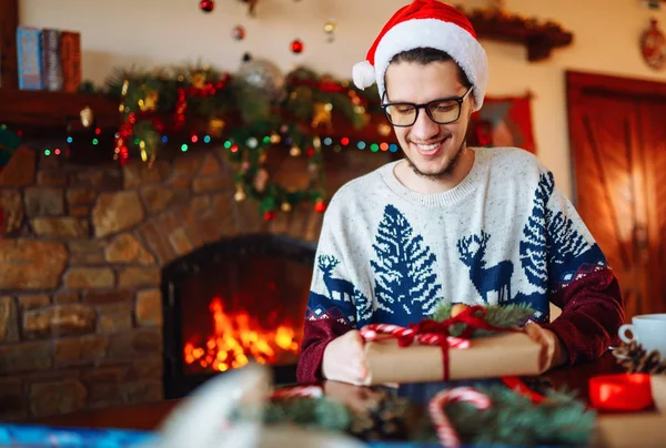 Happy man in a Santa hat creates gifts. The man made them himself and is going to give for the holidays. Concept of gifts and greetings for Christmas and New Year. Beautiful background with fireplace.