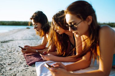 Sahilde telefonu olan üç kız. Genç kızlar güneşlenir ve fotoğraf çekilir. Plaj tatili ve yaz tatili konsepti.