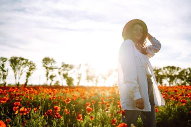 Şapkalı genç kız haşhaş tarlasında poz veriyor. Gün batımında haşhaş tarlasında yürüyen kıvırcık kız portresi. Yaz zamanı..