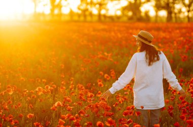 Şapkalı genç kız haşhaş tarlasında poz veriyor. Gün batımında haşhaş tarlasında yürüyen kıvırcık kız portresi. Yaz zamanı..