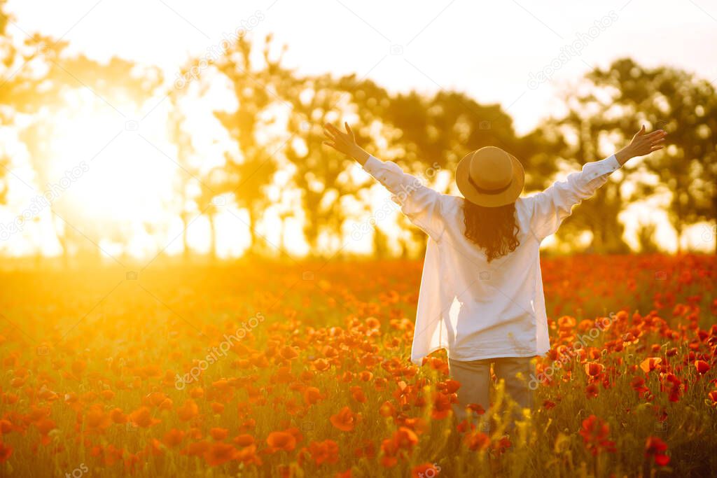 Young girl in hat posing in the poppy field. Portrait of curly girl walking in poppy field at sunset. Summertime.