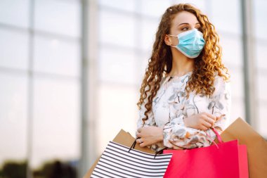 Alışveriş yapan bir kadın. Yüzünde koruyucu steril tıbbi maske takmış alışveriş torbalarıyla gezen genç bir kız. Moda, güzellik ve alışveriş endüstrisinde virüs koruması kavramı.