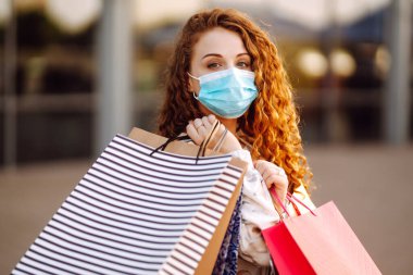 Alışveriş yapan bir kadın. Yüzünde koruyucu steril tıbbi maske takmış alışveriş torbalarıyla gezen genç bir kız. Moda, güzellik ve alışveriş endüstrisinde virüs koruması kavramı.