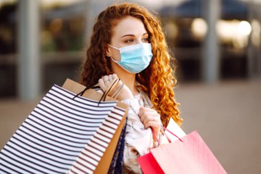 Alışveriş yapan bir kadın. Yüzünde koruyucu steril tıbbi maske takmış alışveriş torbalarıyla gezen genç bir kız. Moda, güzellik ve alışveriş endüstrisinde virüs koruması kavramı.