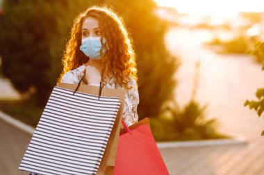 Alışveriş yapan bir kadın. Yüzünde koruyucu steril tıbbi maske takmış alışveriş torbalarıyla gezen genç bir kız. Moda, güzellik ve alışveriş endüstrisinde virüs koruması kavramı.
