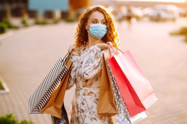 Alışveriş yapan bir kadın. Yüzünde koruyucu steril tıbbi maske takmış alışveriş torbalarıyla gezen genç bir kız. Moda, güzellik ve alışveriş endüstrisinde virüs koruması kavramı.