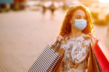 Alışveriş yapan bir kadın. Yüzünde koruyucu steril tıbbi maske takmış alışveriş torbalarıyla gezen genç bir kız. Moda, güzellik ve alışveriş endüstrisinde virüs koruması kavramı.