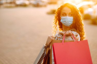 Alışveriş yapan bir kadın. Yüzünde koruyucu steril tıbbi maske takmış alışveriş torbalarıyla gezen genç bir kız. Moda, güzellik ve alışveriş endüstrisinde virüs koruması kavramı.