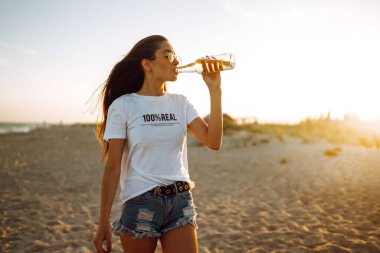 Yaz tatilinde gün batımında sahilde bir şişeden bira içen genç bir kız. Yaz tatili, rahatlama ve yaşam tarzı konsepti.