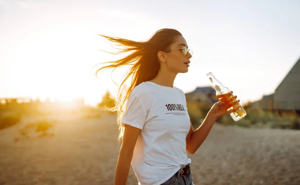 Young girl drinking beer from a bottle on the beach at sunset during  summer vacation. Summer holidays, relax and lifestyle concept.