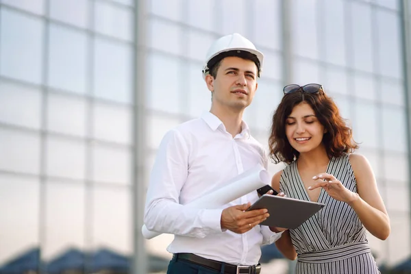 Two workers in a helmet at a work object discuss working issues. Man and woman at the construction site discuss construction project. Business, building, paperwork and people concept.