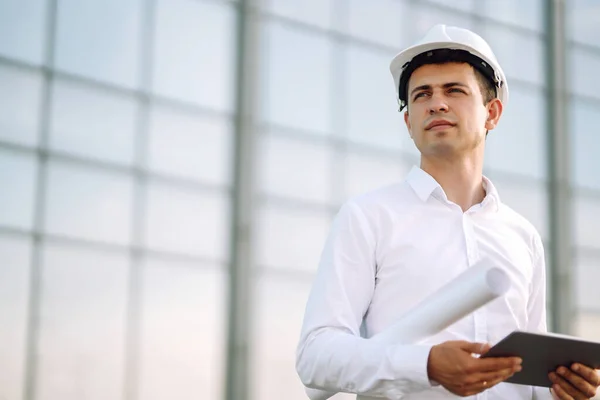 Young worker in a helmet and with a tablet at a work object. Male engineer at a construction site with a tablet computer. Business, building, paperwork and people concept.