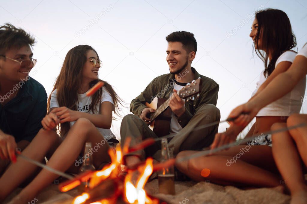 Group of happy friends frying sausages on campfire at the beach. A company of young people came together for a barbecue.
