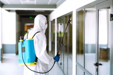 COVID-19 koronavirüs için müteahhit dezenfekte ofisi. Koruyucu giysili ve maskeli bir dezenfektan ofiste dezenfektan sıkar. Yüzeyleri olan zatürree virüsünün yayılmasını önlemek.