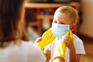 Sarı eldivenli anne, bebek steril tıbbi maskesini takıyor. Salgının yayılmasını önleme ve koronavirüsü tedavi etme kavramı, karantina şehrinde salgın. Covid - 19.