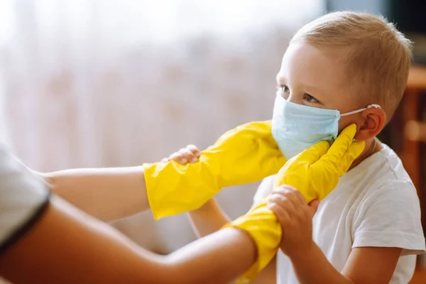 Mother in yellow gloves  puts on her baby sterile medical mask. The concept of preventing the spread of the epidemic and treating coronavirus, pandemic in quarantine city. Covid -19.