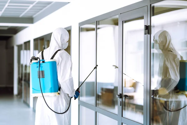 Contractor in a protective suit and mask sprays disinfectants in office before work. Protection against COVID-19 disease. Prevention of spreding pneumonia virus with surfaces.