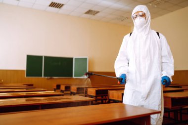 COVID-19 'u önlemek için sınıf dezenfekte ediliyor. Koronavirüsün yayılmasını önlemek için kimyasal sprey kullanan koruyucu koruyucu koruyucu koruyucu giysili bir adam, karantina şehrinde salgın.. 