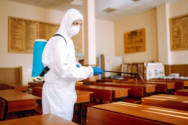 Disinfecting school class to prevent COVID-19. Man in protective hazmat suit with spray chemicals to preventing the spread of coronavirus, pandemic in quarantine city.