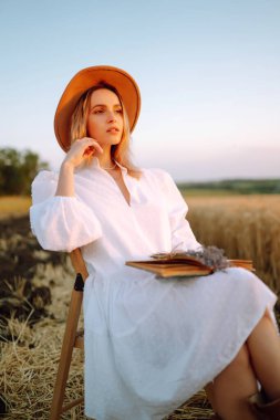 Beyaz elbiseli şık bir kız ve buğday tarlasında kitabı olan bir şapka. Gün batımında mutlu genç bir kadın ya da yaz doğasında gün doğumunda. Moda ve yaşam tarzı konsepti.