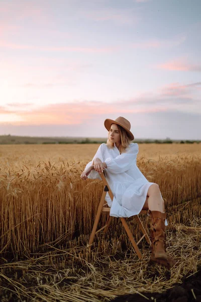 Stylish young girl in a summer white dress and hat posing in a golden wheat field. Fashion, glamour, lifestyle concept.