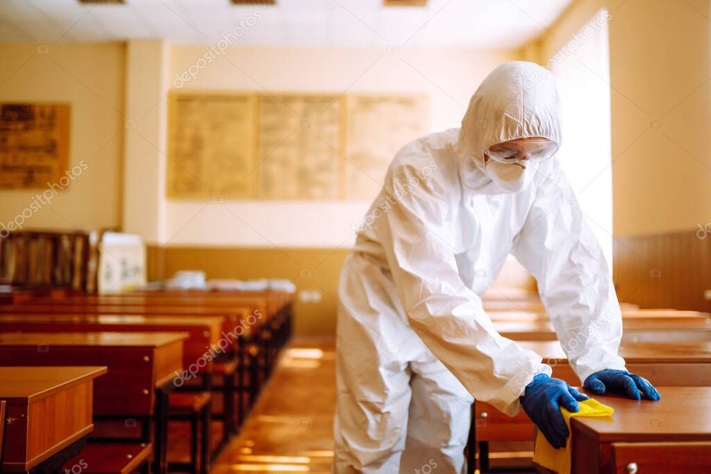 Man in protective hazmat suit washes school desk during coronavirus pandemic. COVID-19.