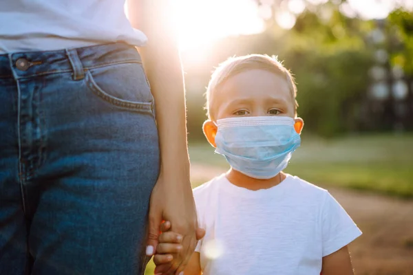 Little boy in sterile medical mask walks by the hand with mom in the park. Together during pandemic. Covid-2019.