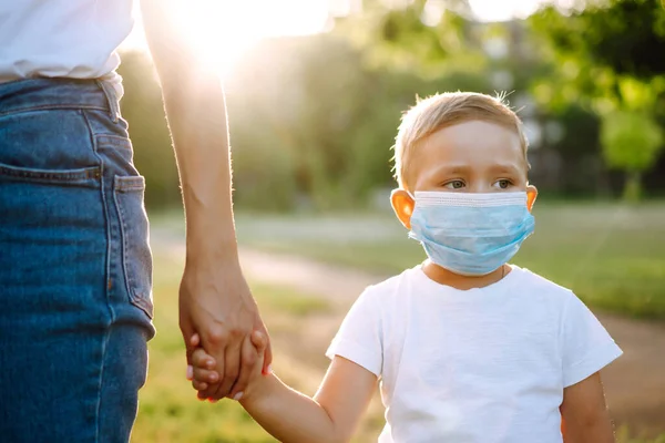 Little boy in sterile medical mask walks by the hand with mom in the park. Together during pandemic. Covid-2019.