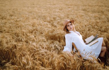 Beyaz keten elbiseli ve şapkalı genç bir kadın altın buğday tarlasında güneşli bir günün tadını çıkarıyor. Yaz, güzellik, moda, cazibe, yaşam tarzı konsepti.