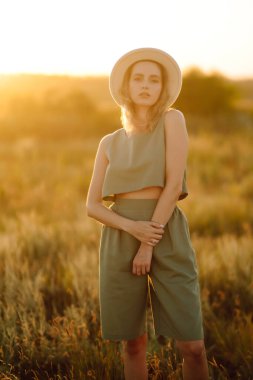 Genç ve güzel bir kadın gün batımında doğada vakit geçiriyor. Altın buğday tarlasında güneşli bir günün tadını çıkaran şık bir kız. Yaz, güzellik, moda, cazibe, yaşam tarzı konsepti.