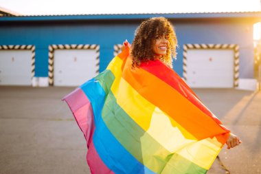 Gün batımında LGBT gökkuşağı bayrağı taşıyan güzel Afro-Amerikan lezbiyen kız. Igbt gurur bayrağıyla poz veren kıvırcık saçlı genç kadın..