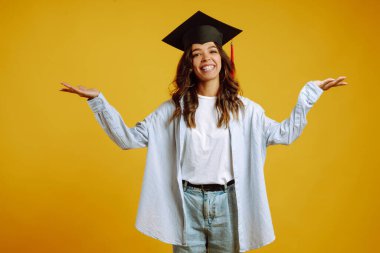 Kafasında mezuniyet şapkasıyla sarı arka planda poz veren Afrikalı Amerikalı bir kızın portresi. Mezuniyet, üniversite, üniversite, uzaktan eğitim kavramı.