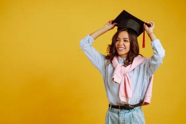 Kafasında mezuniyet şapkasıyla sarı arka planda poz veren Afrikalı Amerikalı bir kızın portresi. Mezuniyet, üniversite, üniversite, uzaktan eğitim kavramı.