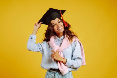 Kafasında mezuniyet şapkasıyla sarı arka planda poz veren Afrikalı Amerikalı bir kızın portresi. Mezuniyet, üniversite, üniversite, uzaktan eğitim kavramı.