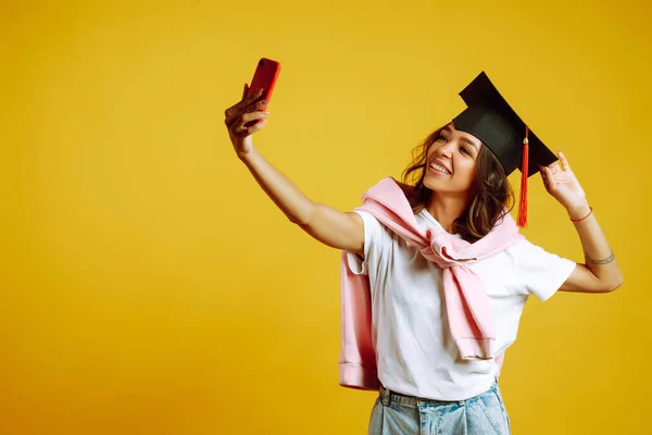 African American girl in a graduation hat on her head take selfie on a yellow background. Graduation, university, college, distance education concept.