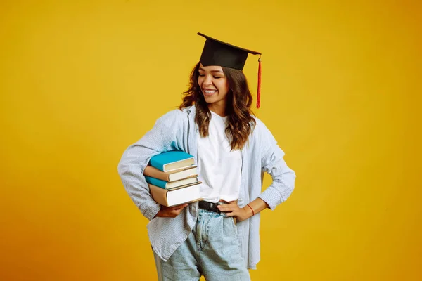 頭には卒業帽子を被った大学院生の女の子がいて 本は黄色い背景に立っています アフリカ系アメリカ人の少女が笑顔でポーズ 遠隔教育の概念 — ストック写真